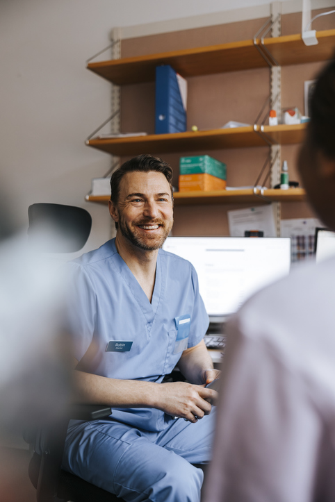 Happy male doctor in blue scrubs talking with patient while sitting in medical clinic room